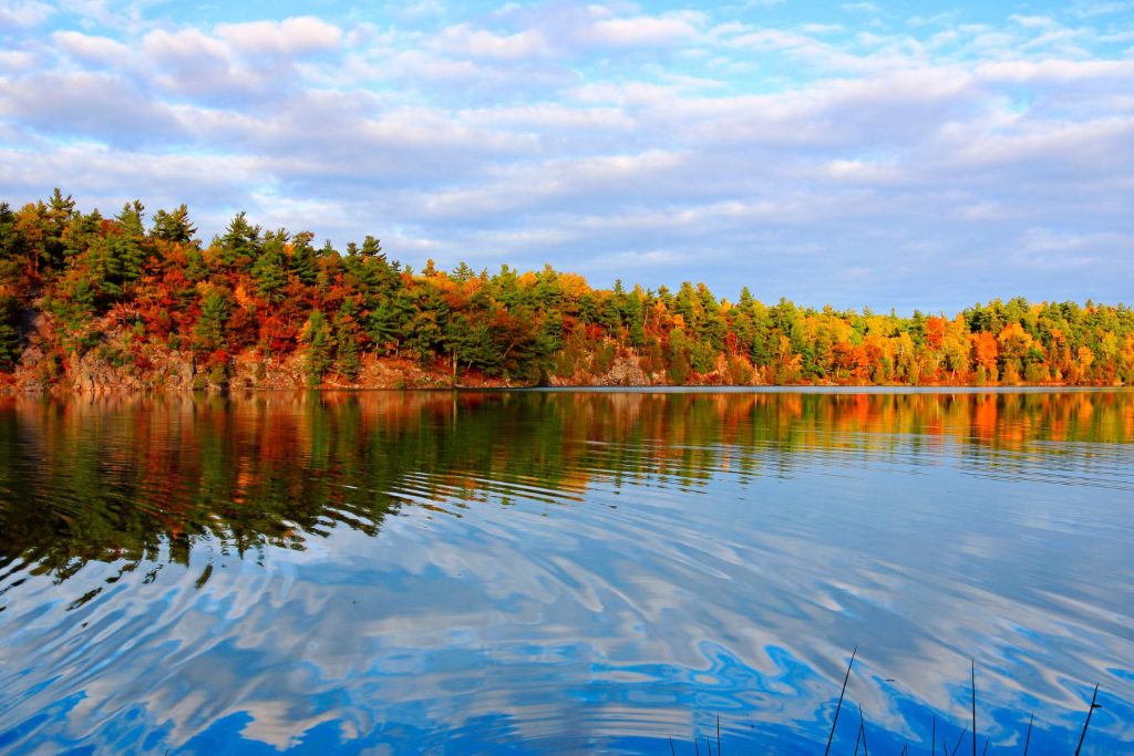 Beautiful fall colour landscape with reflections of forest in greens, red, orange and yellow leaves, and rocky shoreline and clouds on the water along Pink Lake, Gatineau Park. A provincial park near the shore of the Ottawa River in Quebec, Canada.