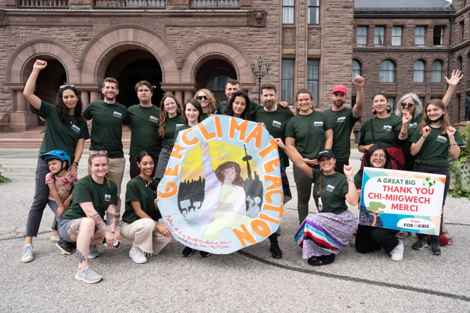 A group of young people stand together holding a large round sign that says Gen Climate Action outside Queen’s Park in Toronto. They are wearing dark green t-shirts. Some are standing with their arms raised, some are kneeling. A second signs says, “A great big thank you.”